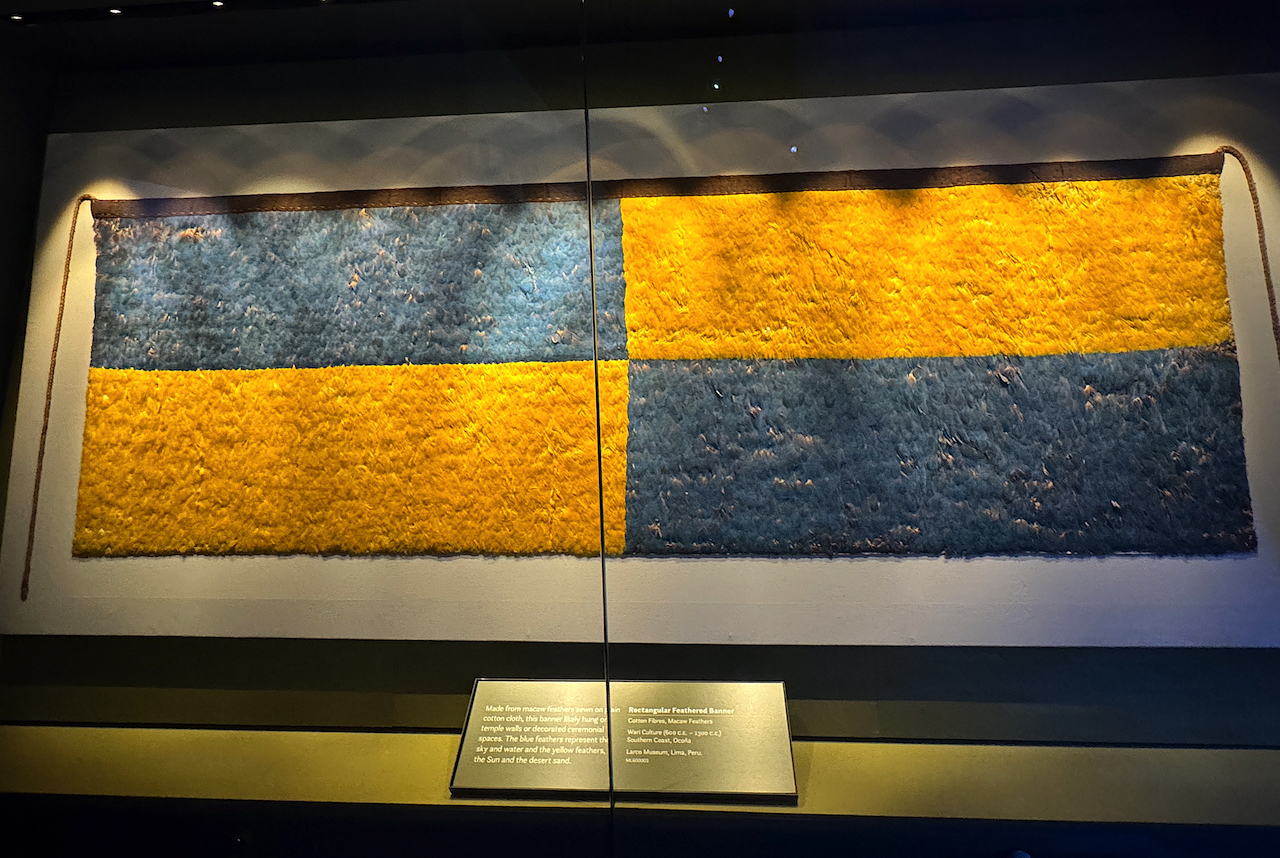 Banner made from macaw feathers sewn on cotton, blue representing the sky and water, yellow the sun and desert sand.